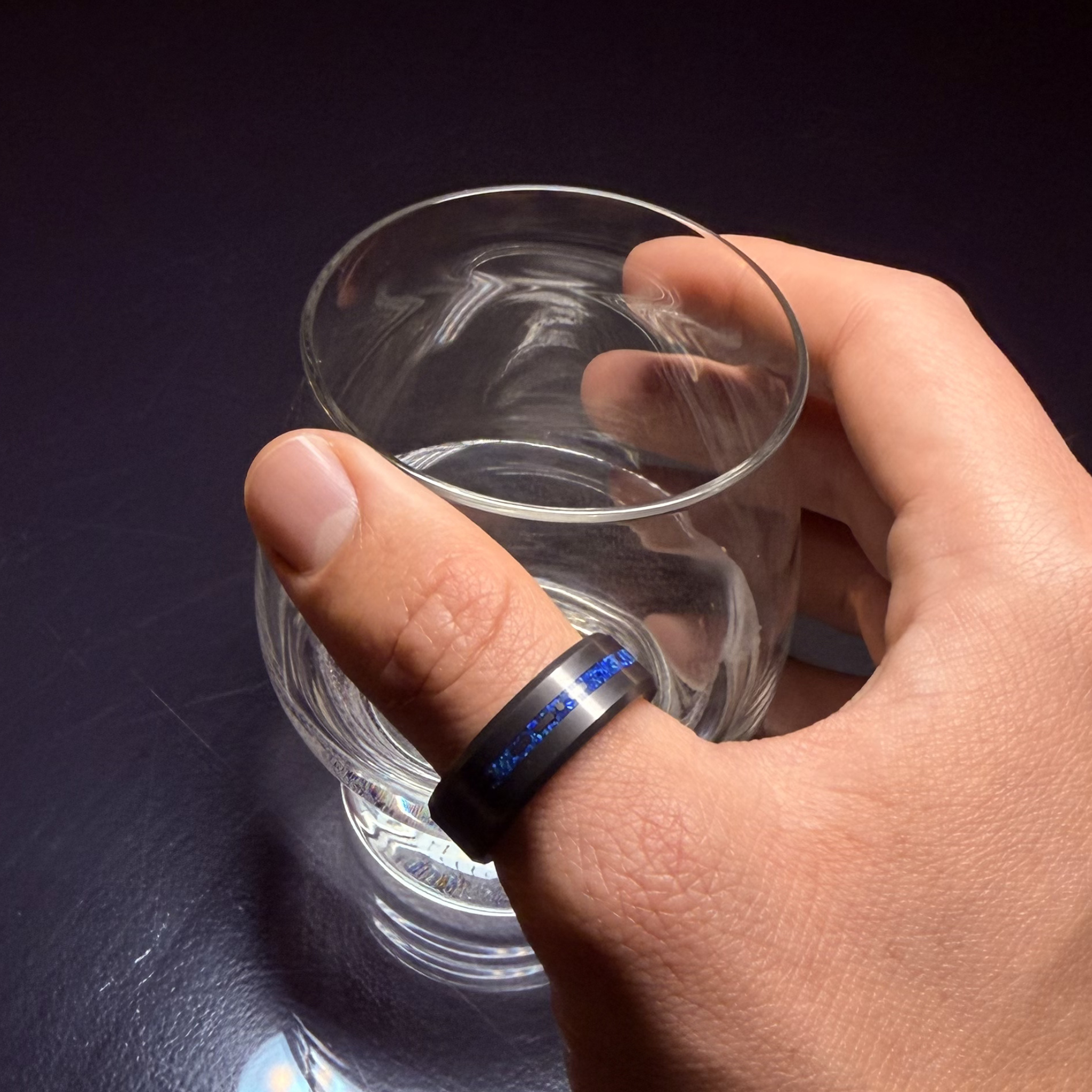 Hand wearing Solid Black Diamond ring with a laser engraved inlay reading "I love you" in morse code, filled with dark blue opal inlay holding a glass on a reflective background.