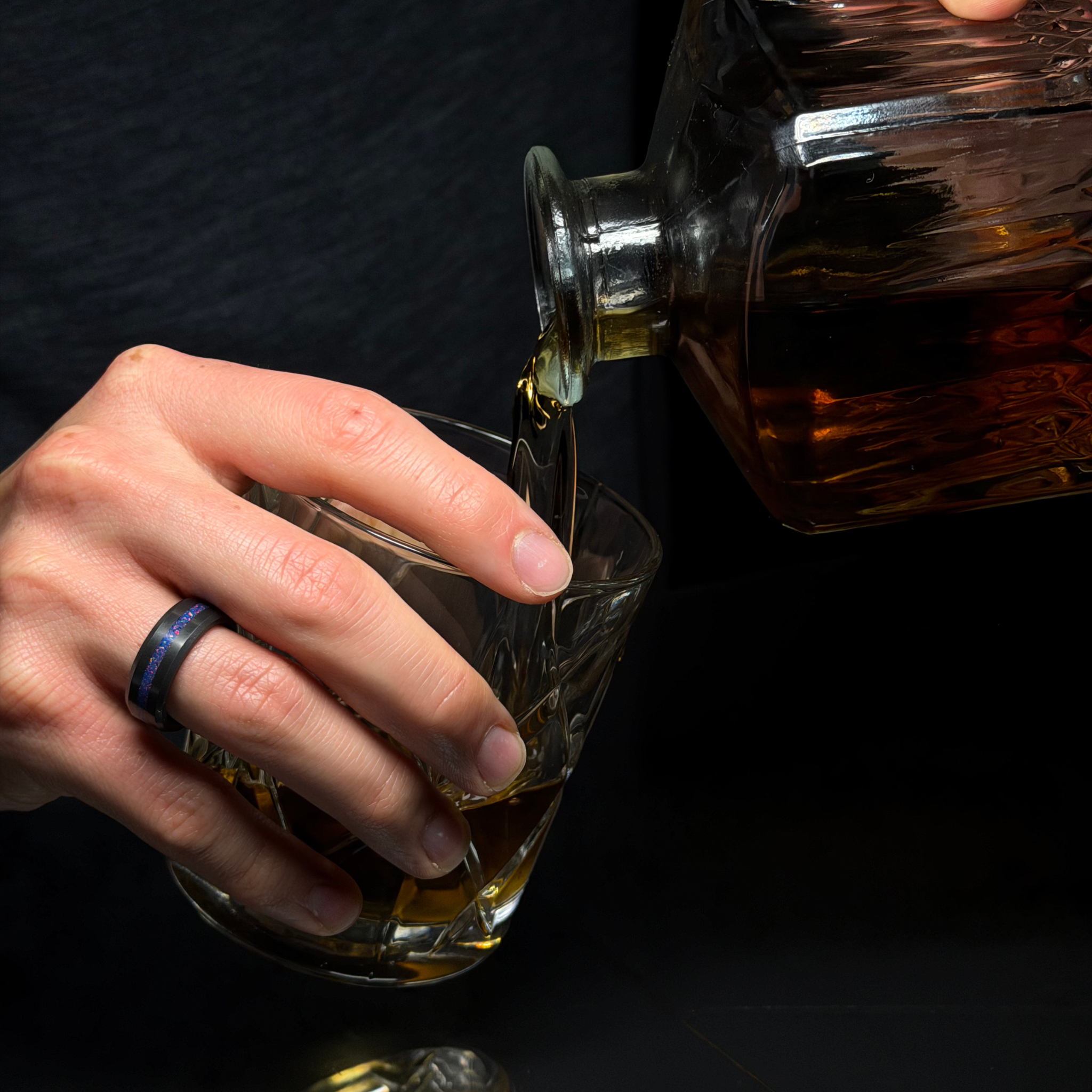 Hand wearing solid black diamond ring with a purple opal inlay pouring whiskey from a bottle into a glass against a dark background