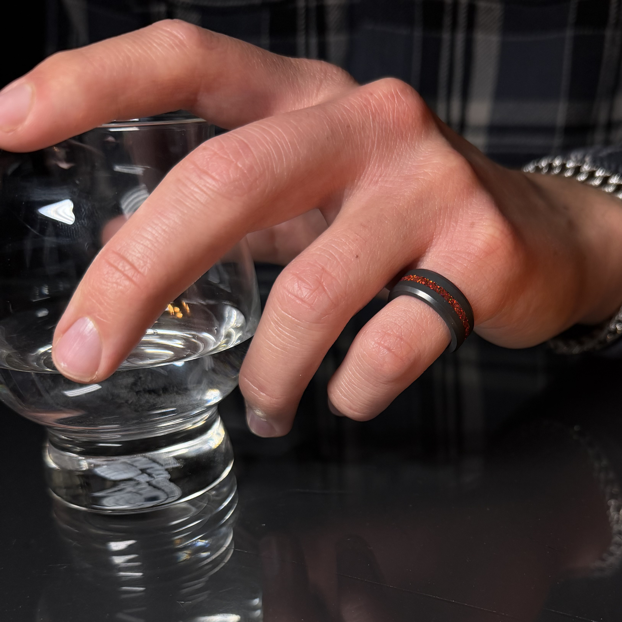 Hand holding a clear glass wearing a solid black diamond ring with a red opal inlay on a reflective surface