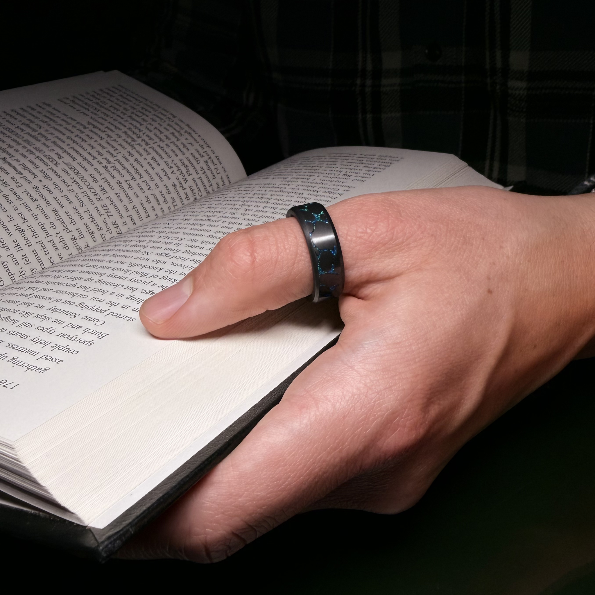 Hand holding a book and wearing a Solid Black Diamond ring with laser engraved river stone inlay filled with blue opal 